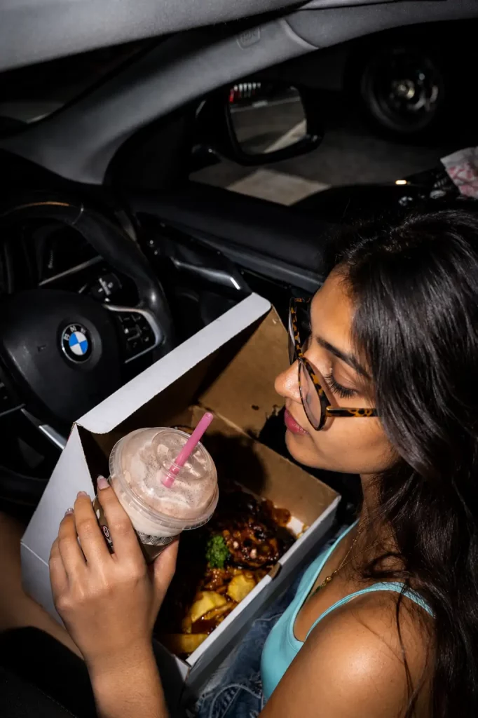 Young Indian woman sitting in a BMW at night, holding a milkshake with a pink straw and enjoying a Burger Box takeaway meal in the car.